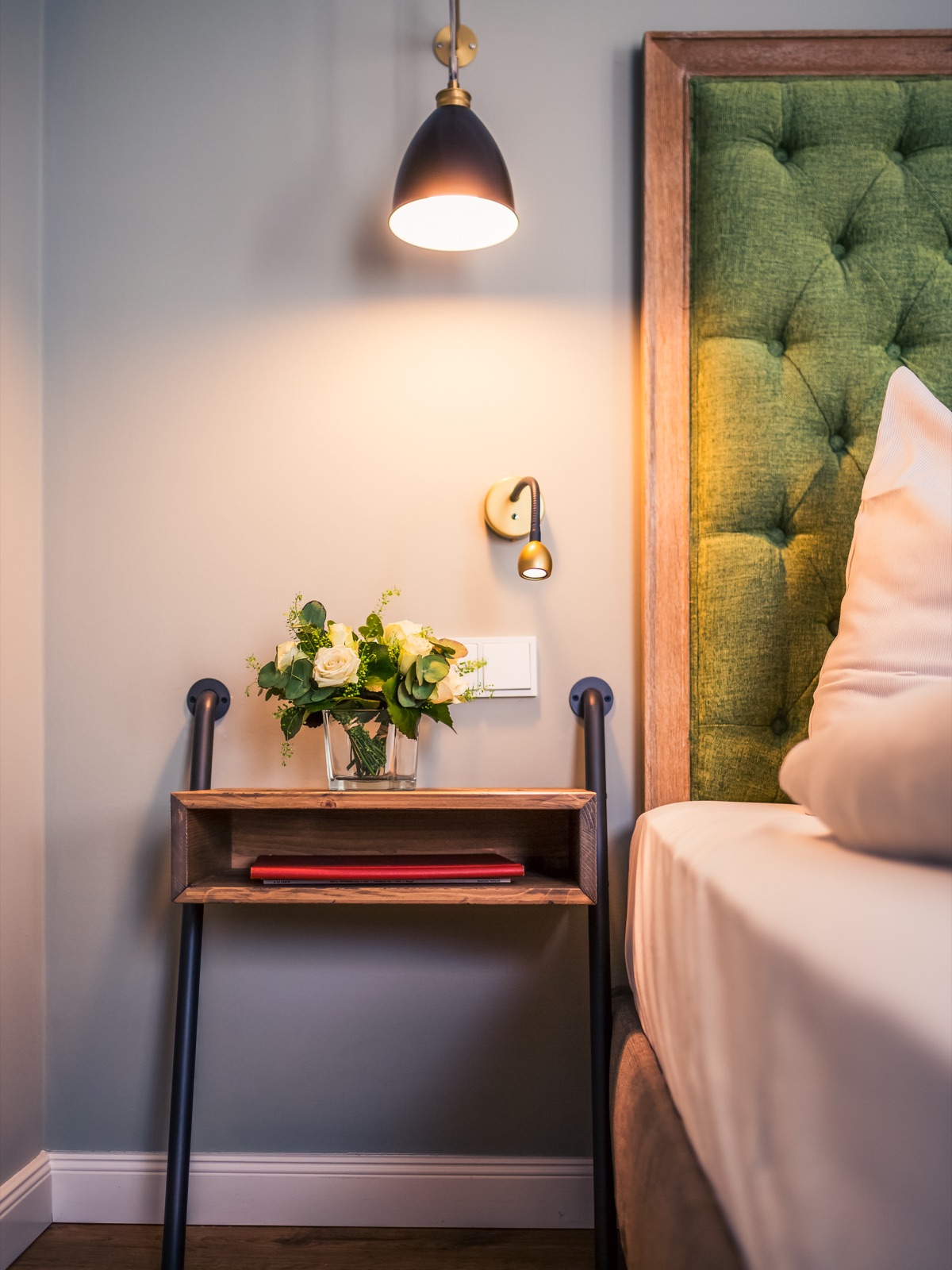 Hotelzimmer auf Gut Altona – Bett mit grünem Polsterkopfteil, Leselampe und Nachttisch mit frischen Blumen